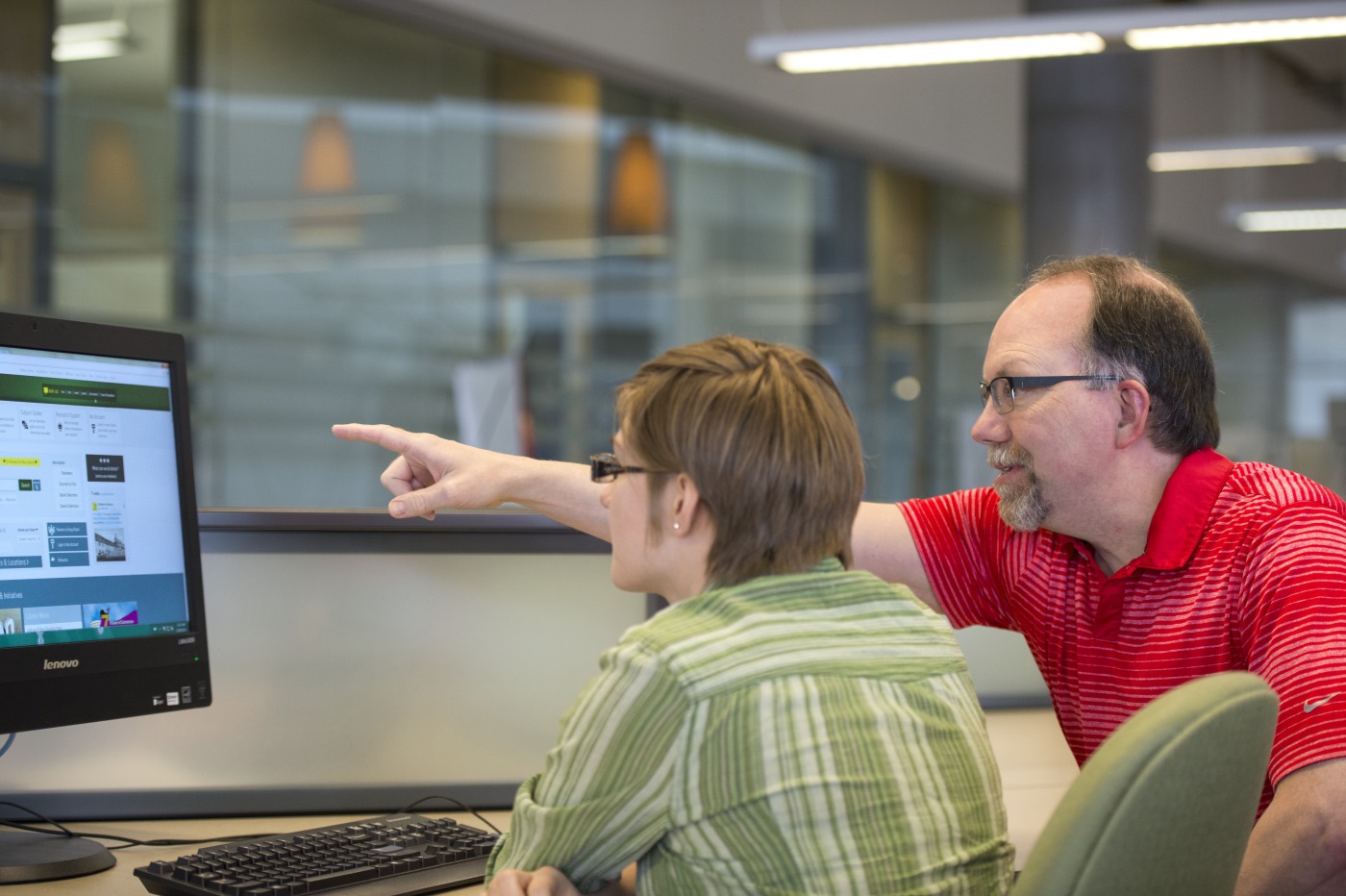 Student sitting at a computer desk with the instructor leaning and pointing at the scren