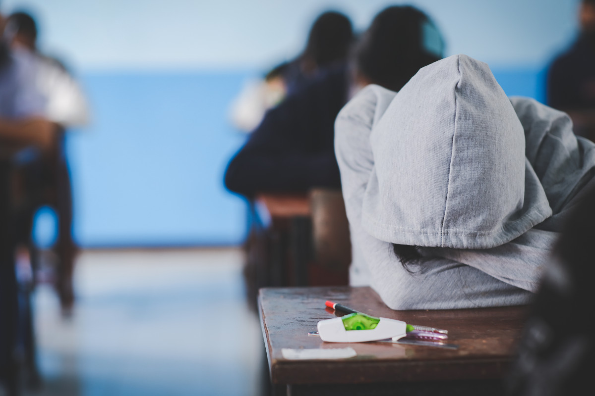 A student sits with their head facedown on a desk, a grey hoodie pulled up over their head.