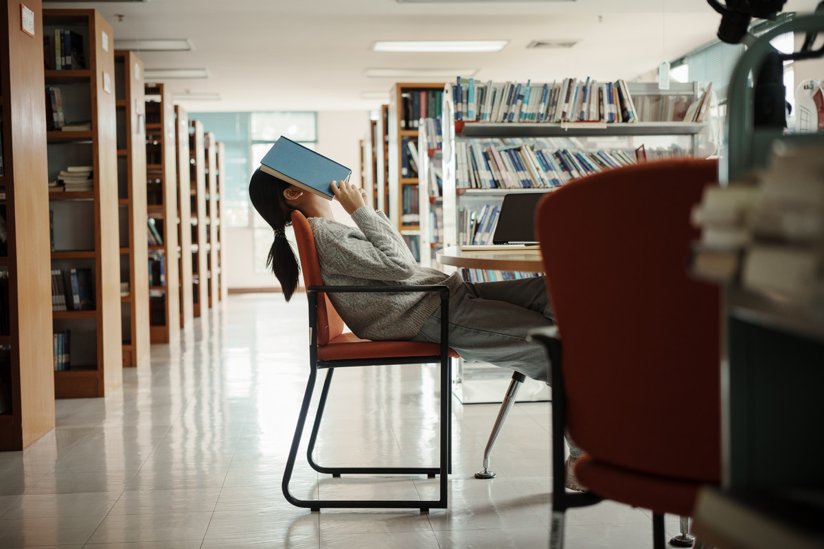 A student sits in a library with a book held over their face, appearing exhausted.