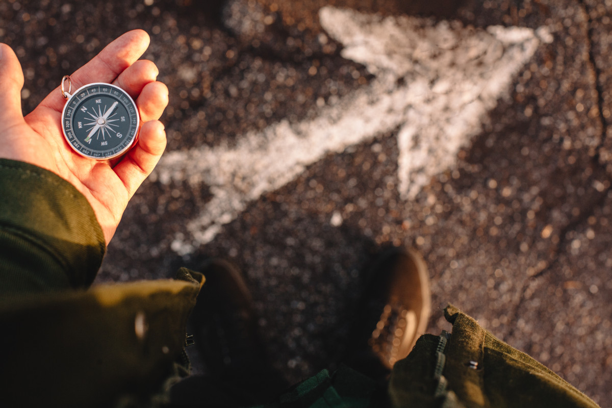 A person holds a compass in their hand, looking down at their feet — where an arrow is painted on the ground.